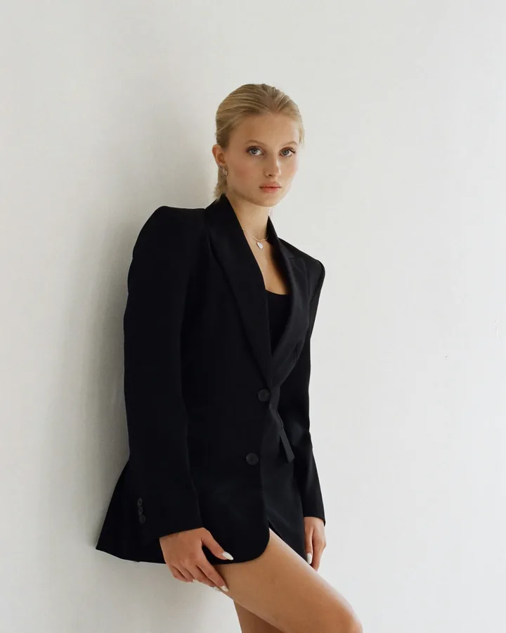 Femme en blazer noir adossée à un mur blanc