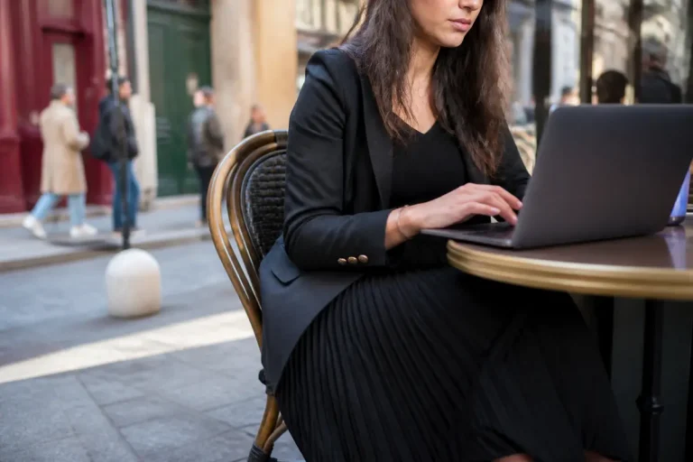 Femme travaillant sur ordinateur, look élégant avec jupe longue plissée noire en terrasse.