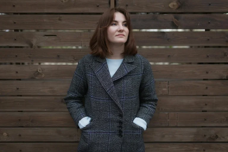 Femme en manteau de laine, les mains dans les poches, devant un mur en bois